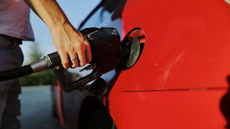 Close-up of a person's hand holding a fuel nozzle while refueling a red car at an outdoor gas station. Featured image for Kim Monson Newsroom: Iran war's oil shock reaches metro Denver kitchen tables and closing tables